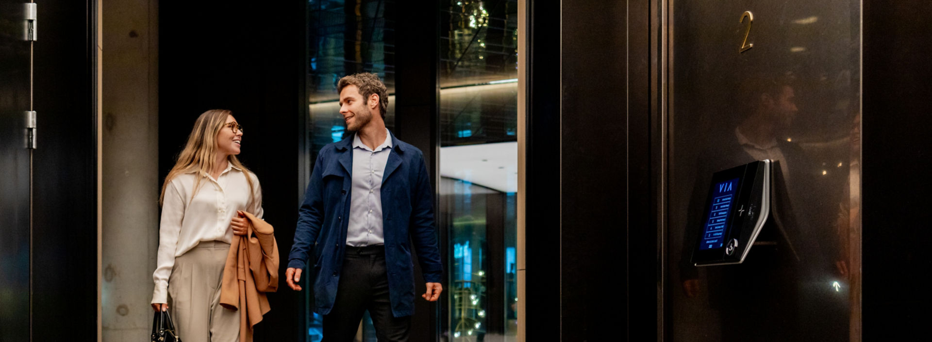 A man and a woman in casual business wear walk towards an elevator while talking happily.